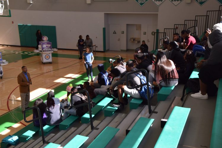 Students gather on gymnasium bleachers to listen to a presenter.